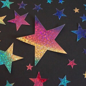Könnte beinhalten: Eine Ansammlung verschieden großer, schillernder Sternaufkleber auf schwarzem Hintergrund. Die Sterne schimmern in Farben wie Gold, Rosa, Blau und Grün und erzeugen ein himmlisches Thema. Ideal für Bastelarbeiten und Dekorationen.