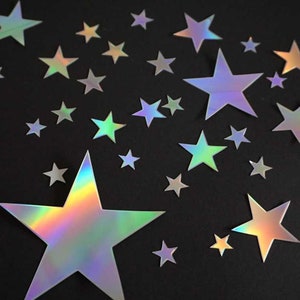 Könnte beinhalten: Eine Ansammlung holografischer sternförmiger Aufkleber in verschiedenen Größen auf schwarzem Hintergrund. Die Aufkleber reflektieren ein Regenbogenspektrum an Farben, darunter Grün, Blau und Gelb. Die Sterne variieren in der Größe.