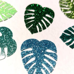 May include: Close-up of several glittery monstera leaf cutouts in shades of green and blue. The leaves have a textured, sparkling appearance and are arranged on a white surface. These decorative elements are ideal for crafting or party decorations.