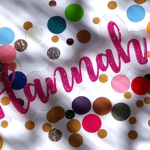 Könnte beinhalten: Ein klarer Aufkleber mit dem Namen "Hannah" in rosa Glitzerschrift, umgeben von bunten Konfetti und kleinen, runden, glitzernden Akzenten. Das Konfetti umfasst Gold, Silber und verschiedene Farben wie Blau, Grün und Orange.