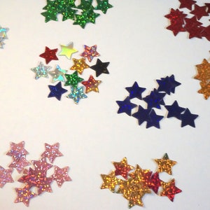 Könnte beinhalten: Eine Sammlung von sternförmigen Pailletten in verschiedenen Farben, darunter Silber, Rot, Grün, Blau, Gold und Rosa. Die Pailletten haben ein holografisches, glitzerndes Finish und sind auf einer weißen Oberfläche verstreut. Ideal für Bastelarbeiten.