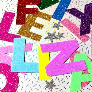 Könnte beinhalten: Glitzerbuchstaben, die die Namen "ELIZA" und "LEXM" in verschiedenen Farben wie Pink, Rot, Gold, Blau, Lila und Grün schreiben. Silberne Glitzersterne sind ebenfalls verstreut.