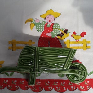 May include: A green ceramic wheelbarrow planter with a wooden plank design. The wheelbarrow is sitting on a white tablecloth with a red and white floral border.