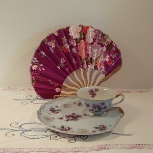 May include: A delicate teacup and saucer set with a floral pattern. The cup and saucer are white with a light blue background and purple flowers. The saucer has a scalloped edge. A purple and pink floral fan is in the background.
