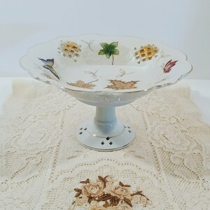 May include: White porcelain pedestal bowl with a scalloped edge and a delicate floral design. The bowl features a lattice pattern and gold accents. The bowl is on a white lace tablecloth.