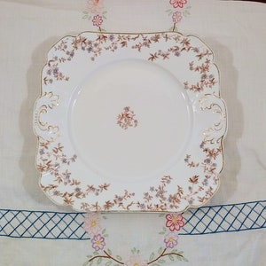 May include: A white square ceramic serving plate with a gold rim and a floral pattern in shades of brown and purple.
