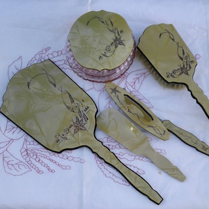 Può includere: Un set da toeletta vintage con un design verde e nero. Il set include uno specchio da mano, una spazzola per capelli, un pettine e un barattolo di polvere con un coperchio. Il set ha un design floreale con un contorno nero.