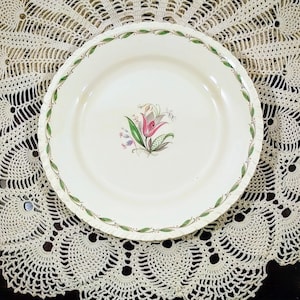 May include: A white ceramic dinner plate with a green and brown floral design featuring a pink tulip. The plate has a scalloped edge and is surrounded by a white crocheted doily.