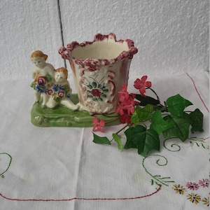 May include: A vintage ceramic planter with two cherub figurines holding flowers. The planter is decorated with floral designs in shades of pink, green, and white. The base of the planter is green and has a scalloped edge.