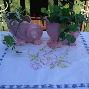 Può includere: Due vasi in ceramica rosa a forma di conchiglia con piante di edera verde al loro interno. I vasi sono appoggiati su una tovaglia bianca ricamata con un motivo floreale.
