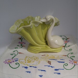 May include: A vintage ceramic swan planter with a green and white glaze. The planter is shaped like a swan with its neck curved up and its head tilted to the side. The planter is sitting on a white tablecloth with embroidered swans.