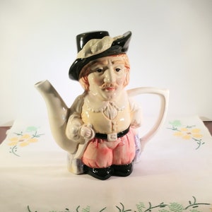 Puede incluir: Una tetera de cerámica con forma de hombre que lleva un sombrero negro, una camisa blanca y pantalones rosas. La tetera tiene un pico y un asa blancos.