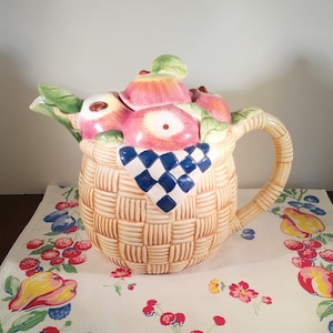 May include: A ceramic teapot shaped like a basket filled with apples. The teapot is decorated with a checkered blue and white cloth and has a brown handle.