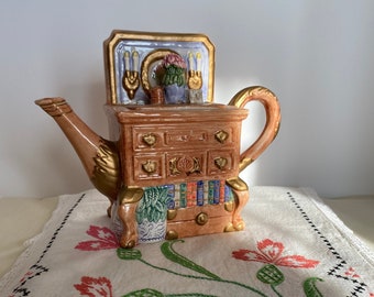 May include: A ceramic teapot shaped like a dresser with a mirror and candles on top. The teapot is painted in shades of brown, gold, and white. It is sitting on a white embroidered tablecloth with pink and green flowers.