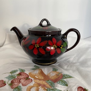 Könnte beinhalten: Eine dunkelbraune Teekanne mit Blumenmuster. Die Teekanne hat rote Blumen mit gelben Zentren und grünen Blättern. Die Teekanne steht auf einer weißen Tischdecke mit einem gestickten Blumenmuster.