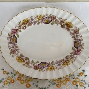 Könnte beinhalten: Eine weiße ovale Servierplatte mit goldfarbenem Rand und einem Blumendesign in Braun-, Gelb- und Lilatönen.