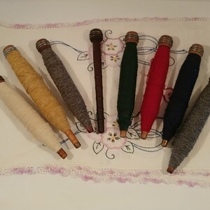 May include: Eight wooden spools of thread, each with a different color of thread wound around it. The spools are arranged in a semi-circle on a white cloth with a pink lace trim.