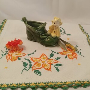 Könnte beinhalten: Ein grüner Keramik-Schuh-Pflanztopf mit einer kleinen gelben Figur eines Zwergs, der oben sitzt. Der Topf steht auf einer weißen Häkeldeckchen mit gestickten Blumen.