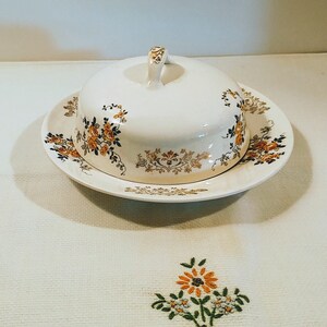 Puede incluir: Un mantequillero de cer&aacute;mica blanca con tapa y un dise&ntilde;o floral en tonos naranja, marr&oacute;n y dorado. El plato est&aacute; sobre un mantel blanco con flores bordadas.