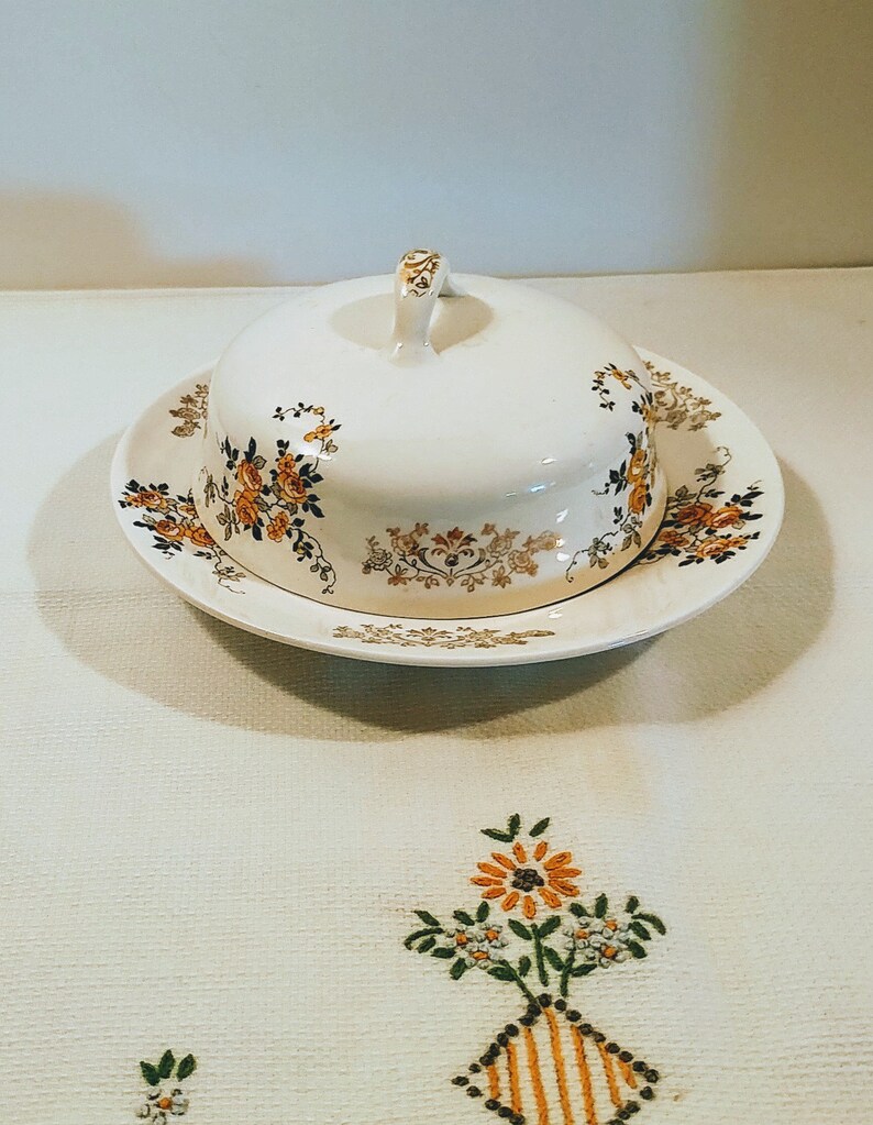 Puede incluir: Un mantequillero de cer&aacute;mica blanca con tapa y un dise&ntilde;o floral en tonos naranja, marr&oacute;n y dorado. El plato est&aacute; sobre un mantel blanco con flores bordadas.