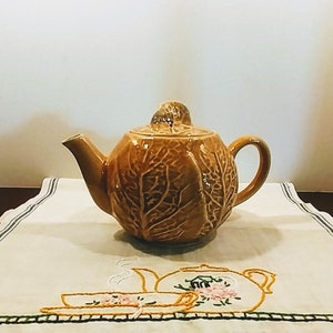 May include: A brown ceramic teapot shaped like a cabbage with a leafy design. The teapot is sitting on a white tablecloth with an embroidered design of a teapot and teacups.