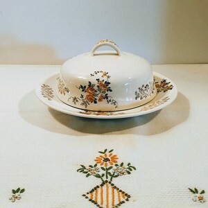 Puede incluir: Un mantequillero vintage blanco con un dise&ntilde;o floral. El mantequillero tiene una tapa con un pomo en la parte superior y un plato a juego.
