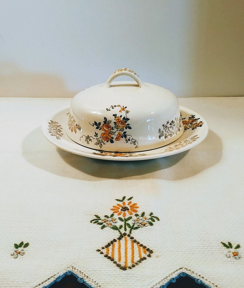 Puede incluir: Un mantequillero vintage blanco con un dise&ntilde;o floral. El mantequillero tiene una tapa con un pomo en la parte superior y un plato a juego.