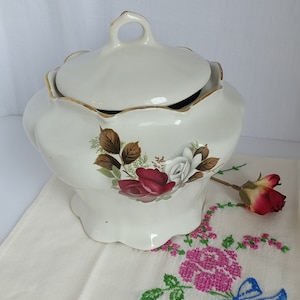 Könnte beinhalten: Weiße Zuckerdose aus Keramik mit Deckel und floralem Design mit roten und weißen Rosen. Die Schale hat einen gewellten Rand und einen goldenen Rand.