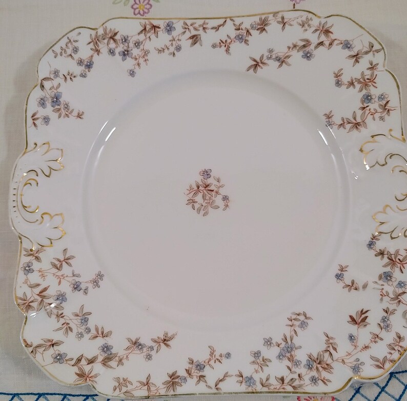 May include: A white square plate with a gold rim and a delicate floral pattern in shades of blue and brown. The plate has a scalloped edge and a small floral design in the center.