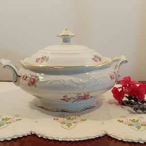 Może przedstawiać: Biała porcelanowa miska do serwowania z pokrywką i kwiatowym wzorem. Miska ma złotą obwódkę i jest ozdobiona różowymi, czerwonymi i żółtymi kwiatami. Miska stoi na białym koronkowej serwetce.