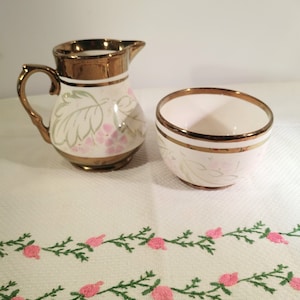 May include: A white ceramic creamer and sugar bowl set with a floral design and copper accents. The creamer has a handle and a spout. The sugar bowl is round and has a wide rim.