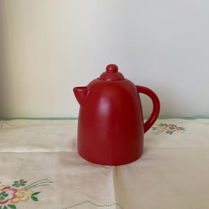 Puede incluir: Una tetera de cerámica roja sólida con un cuerpo redondeado, un asa curva y un pequeño pico. La tapa tiene un pequeño pomo en la parte superior. La tetera está sobre un mantel blanco con bordados florales.