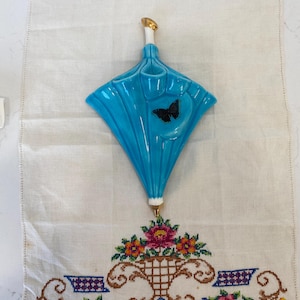 May include: A turquoise ceramic umbrella-shaped wall pocket with a black butterfly accent. The umbrella hangs above a white embroidered table runner with a floral basket design. The umbrella has a gold-colored top.