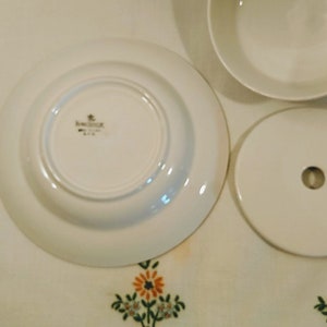 Puede incluir: Plato de cer&aacute;mica blanco con borde dorado y un logotipo dorado que dice "Bavaria" en el centro. El plato est&aacute; sobre un mantel blanco con un patr&oacute;n floral.