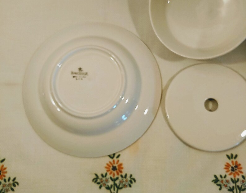 Puede incluir: Plato de cer&aacute;mica blanco con borde dorado y un logotipo dorado que dice "Bavaria" en el centro. El plato est&aacute; sobre un mantel blanco con un patr&oacute;n floral.