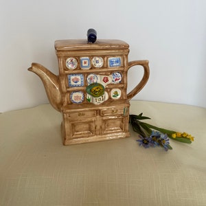 Könnte beinhalten: Eine braune Teekanne aus Keramik in Form eines Miniaturhauses mit einem blauen Knopf auf dem Deckel. Die Teekanne ist mit bunten Fliesen und einem Blumendesign verziert.