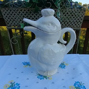 May include: White ceramic pitcher shaped like a duck with a lid. The pitcher is sitting on a white tablecloth with blue and yellow embroidered flowers.