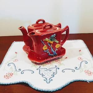 以下が含まれることがあります： 鳥と花のデザインが施された赤い陶器のティーポットセット。セットには小さなティーポットが2つと、それに合うトレイが含まれています。
