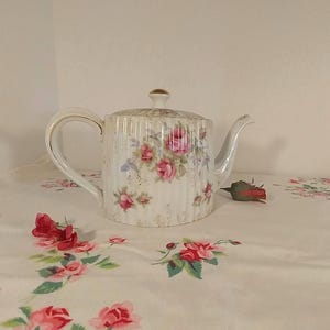 May include: A white porcelain teapot with a floral pattern of pink roses and green leaves. The teapot has a gold trim around the lid and a slightly ribbed design.