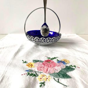 May include: A silver-toned serving dish with a deep blue interior and a decorative, openwork rim. A matching spoon stands upright in the dish's handle. The dish rests on a white embroidered cloth with a floral design in pink, yellow, and blue.