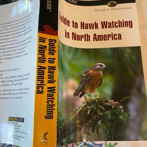 May include: A book titled "Guide to Hawk Watching in North America" by Donald S. Heintzelman. The cover features a hawk perched on a branch. The book's spine is orange with the title and the Falcon Guide logo.