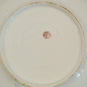 May include: Close-up of the bottom of a white porcelain plate with a red stamp that reads "CARLSBAD A.K. AUS."