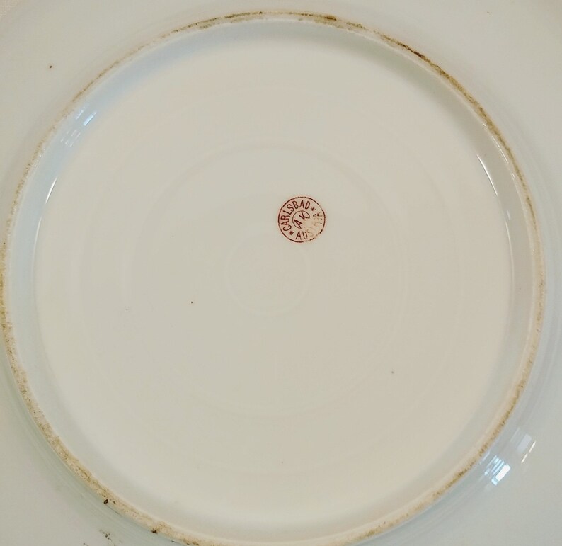 May include: Close-up of the bottom of a white porcelain plate with a red stamp that reads "CARLSBAD A.K. AUS."