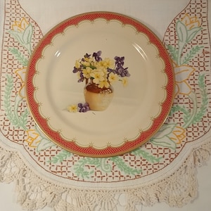 May include: A vintage tin plate with a red and gold rim. The plate features a painted image of a bouquet of purple and yellow flowers in a brown vase.
