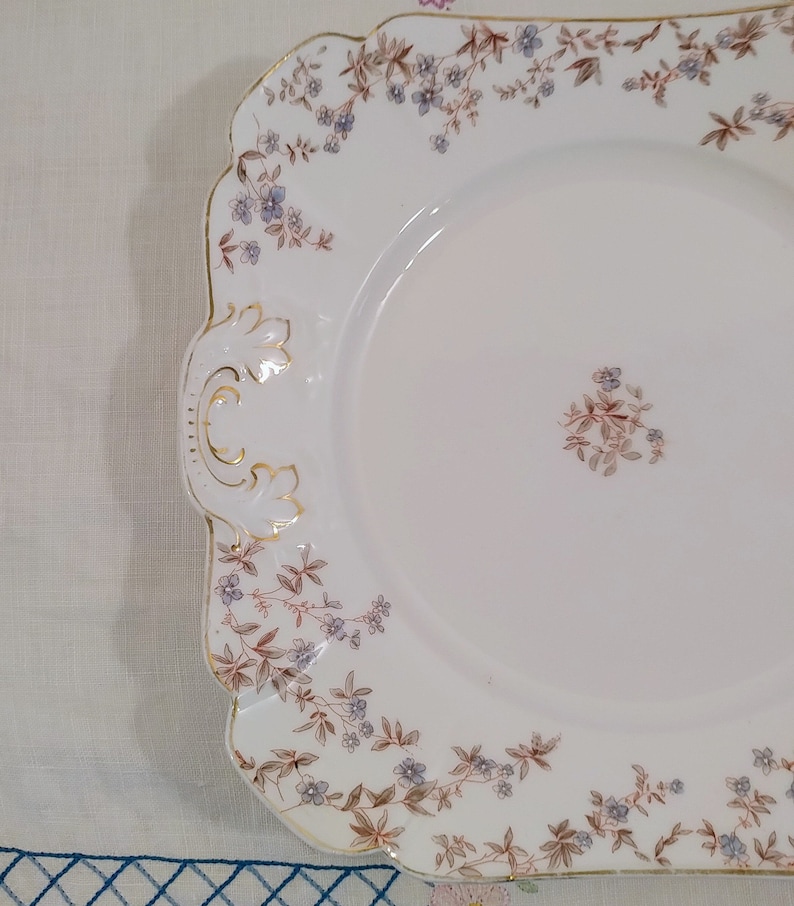 May include: A white porcelain serving platter with a scalloped edge and a floral design in shades of brown and blue. The platter has a gold trim and a raised design on the handle.