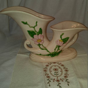 May include: A double-handled, pink ceramic vase with a floral design. The vase features pink flowers with yellow centers and green leaves.