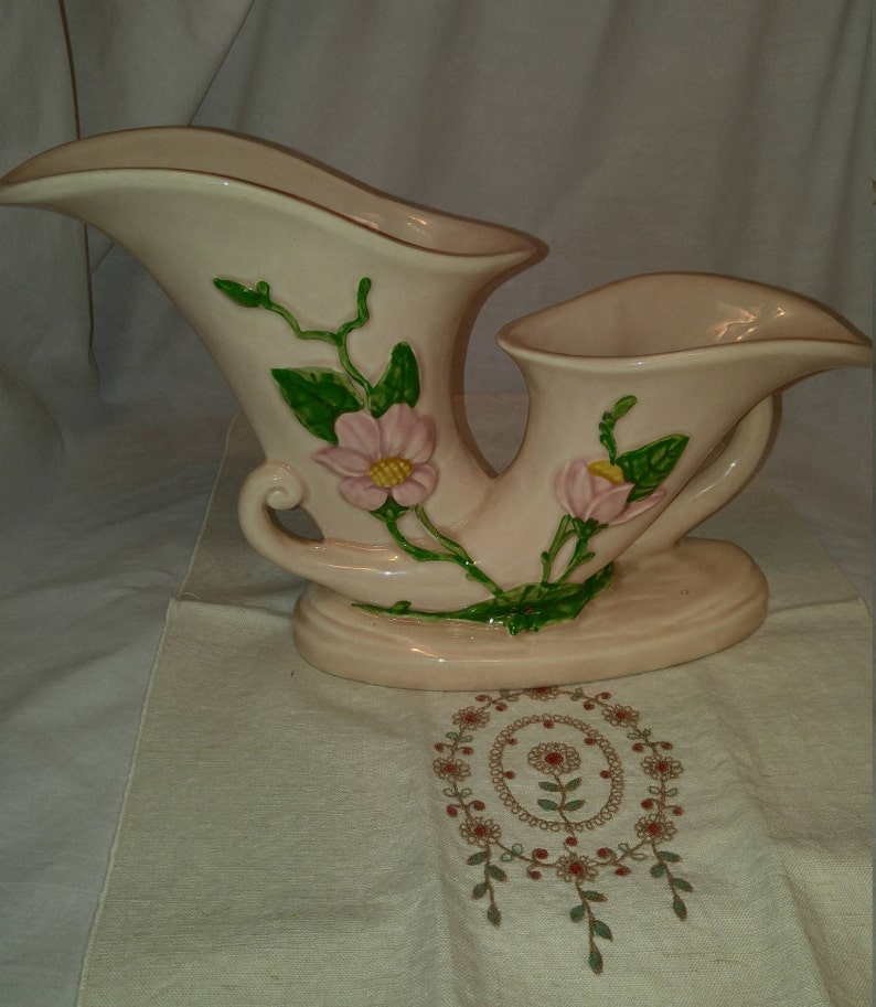 May include: A double-handled, pink ceramic vase with a floral design. The vase features pink flowers with yellow centers and green leaves.