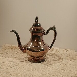 May include: A silver teapot with a rounded body and a decorative handle. The teapot has a shiny finish and a detailed design on the spout.