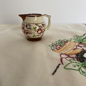 May include: A small white ceramic creamer with a brown and gold floral design. The creamer has a rounded body and a small spout. The handle is attached to the side of the creamer.