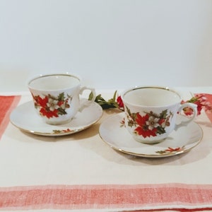 May include: Two white teacups and saucers with a gold rim. The cups and saucers feature a floral pattern with red poinsettia flowers, green leaves, and white accents.